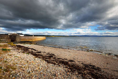An empty promenade to the old lighthouse, gravel beach close-up. Dramatic cloudscape. Ardrishaig,...
