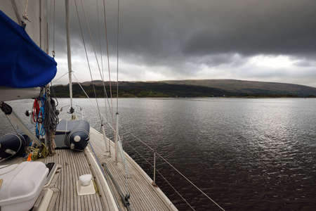 Dark storm sky. Sloop rigged modern yacht with wooden teak deck sailing on a cloudy day. A view from...