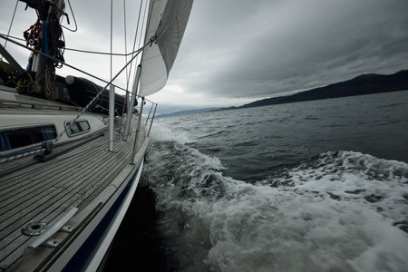 Sloop rigged modern yacht with wooden teak deck sailing on a cloudy day. A view to the mast and sails. Water splashes close-up. Tarbert, Scotland, UKの写真素材