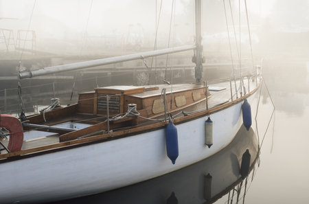 An elegant wooden sailing boat moored to a pier in a thick white morning fog at sunrise. Yacht club...