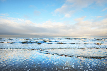 A view from the snow-covered sandy Baltic sea shore at sunset. Riga bay, Latvia. Dramatic sky. Crystal clear blue water. Fickle weather, waves and water splashes. Winter tourism, global warming theme