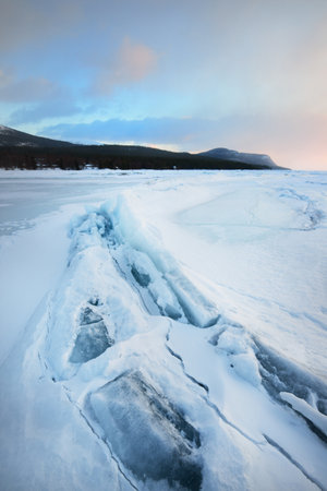 Ice pressure ridge on a frozen lake shore at sunset. Mountain peaks in the background. Colorful cloudscape. Picturesque winter scenery. Ecology, environment, climate change, global warming conceptsの写真素材