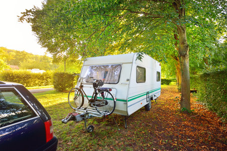 Caravan trailer with a bicycle and a car parked on a green lawn under the trees in a camping site. Normandy, France. Vacations, leisure activity, recreation, tourism, road trip, travel destinationsの写真素材