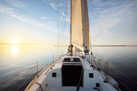 White yacht sailing after the rain at sunset. Close-up view from the deck to the bow. Clear blue sky with glowing clouds reflecting in a still water. Idyllic seascape. Sea, cruise, travel destinationsの写真素材