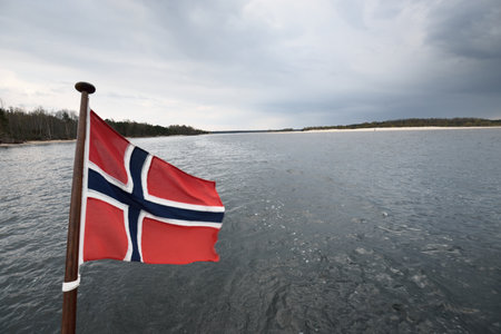 Flag of Norway on the yacht close-up, sailing in Glomma river on a rainy day. Dark storm clouds. Sandy shores and forest in the backgroundの写真素材