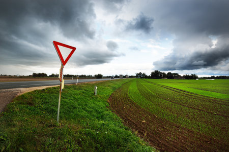 Dark storm clouds above the empty highway through the agricultural fields, road sign close-up. Dramatic evening sky. Latviaの写真素材