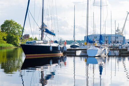 Blue sloop rigged yacht moored to a pier on a clear summer day, close-up. Sailing boats in the background. Port of Riga, Latviaの写真素材