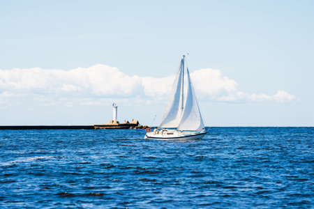 White sloop rigged yacht sailing on a clear day. Lighthouse in the background. Cloudy blue sky. Bay of Riga, Baltic sea, Latviaの写真素材