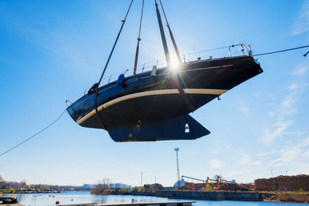 Sailboat lifted by a crane being placed back in the water after winterization in winter. Clear blue...