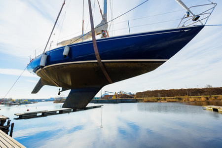 Blue sailboat lifted by a crane being placed back in the water after winterization in winter. Clear...