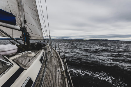 Sloop rigged modern yacht with wooden teak deck sailing near the rocky shores of Tarbert on a cloudy...