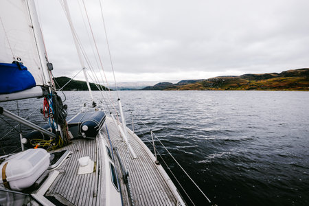 Sloop rigged modern yacht with wooden teak deck sailing near the rocky shores of Kyles of Bute on a cloudy day. Scotland, UK. Close-up view from the deck to the bow, mast and sailsの写真素材