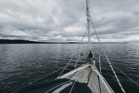 Sloop rigged modern yacht with wooden teak deck sailing near the rocky shores of Tarbert on a cloudy day. Scotland, UK. Close-up view from the deck to the bow, mast and sails. Travel destinationsの写真素材