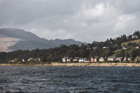 Village on the rocky shore of the Firth of Clyde river, panoramic view from a sailing boat. Dramatic...