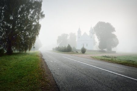 An old white traditional catholic church in a thick mysterious fog, close-up. Travel destinations, sightseeing, national landmark, culture and religion, catholicism, christianity, past, architectureの写真素材