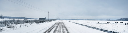 Panoramic view of road through the snow-covered field after a blizzard. Water canal, haystacks. Electricity line, transformer poles. Dark stormy sky. Off-road, logistics, remote village, winterの写真素材