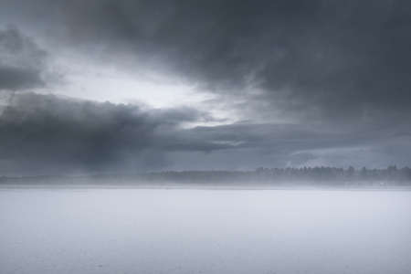 Snow-covered agricultural plowed field and forest under dramatic dark clouds before snowstorm. Winter rural scene. Nature, ecology, environment, climate change, global warmingの写真素材