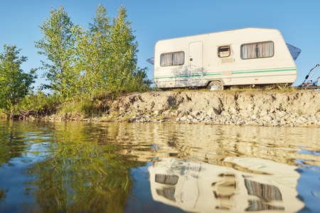 White caravan trailer parked on the rocky river shore on a sunny day. Clear blue sky. Ba; tic...