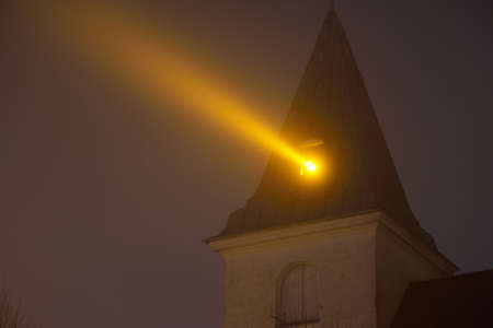 An old historical lighthouse church at night. Latvia. Ray of golden light against clear twilight sky. Dark urban landscape. Landmark, sightseeing, travel destinations. Symbol of hope and peaceの写真素材