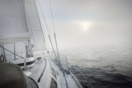 White yacht sailing in the Baltic sea. Thick fog. Sunrise. Sweden. Close-up view from the deck to the bow and sails. Morning sun, glowing clouds. Sport, recreation, leisure activity, cruise, regattaの写真素材