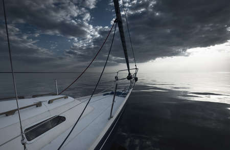 White yacht sailing in the Baltic at sunset. View from the deck to the bow. Glowing clouds, reflections in a still water, natural mirror, Cold silver color. Transportation, cruise, recreation, regattaの写真素材