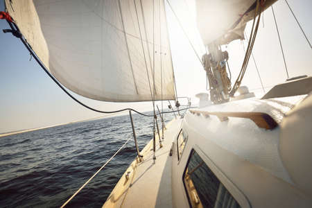 White sloop rigged yacht sailing in an open sea at sunset. Clear sky. A view from the deck to the bow, mast, sails. Transportation, travel, cruise, sport, recreation, leisure activity, racing, regattaの写真素材