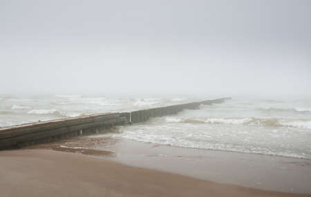 Baltic sea in a thick white fog. An old pier (jetty). Waves, splashing water, storm. Natural textures. Picturesque panoramic monochrome scenery, seascape. Nature, environment, rough weather, dangerの写真素材