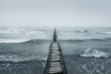 Baltic sea in a thick white fog. An old pier (jetty). Waves, splashing water, storm. Natural textures. Picturesque panoramic monochrome scenery, seascape. Nature, environment, rough weather, dangerの写真素材