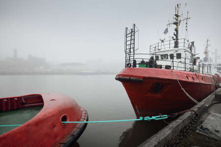 Ships, tugboats and fishing boats (trawlers) moored to a pier in a harbor. Thick white fog. Latvia, Baltic sea. Panoramic view. Service, repair, freight transportation, logistics, industry, commerceの写真素材