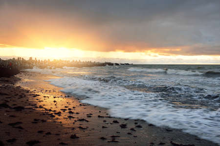 Baltic sea shore (sand dunes) at sunset. Breakwaters and promenade in the background. Soft sunlight, clear sky, glowing clouds, waves. Idyllic seascape. Travel destinations, ecotourism, natureの写真素材
