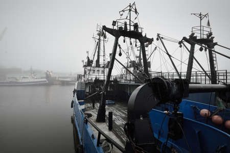 Tugboats and fishing boats (trawlers) moored to a pier in a harbor. Thick white fog. Latvia, Baltic sea. Panoramic view. Service, repair, freight transportation, logistics, industry, commerceの写真素材