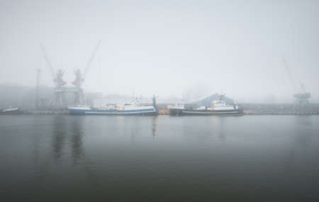 Tugboats and fishing boats (trawlers) moored to a pier in a harbor. Thick white fog. Latvia, Baltic sea. Panoramic view. Service, repair, freight transportation, logistics, industry, commerceの写真素材