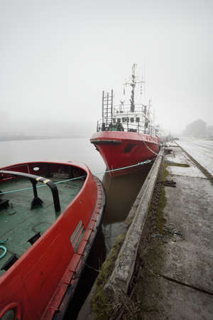 Ships, tugboats and fishing boats (trawlers) moored to a pier in a harbor. Thick white fog. Latvia, Baltic sea. Panoramic view. Service, repair, freight transportation, logistics, industry, commerceの写真素材