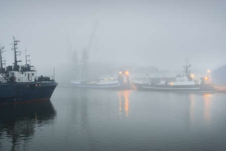 Tugboats and fishing boats (trawlers) moored to a pier in a harbor. Thick white fog. Latvia, Baltic sea. Panoramic view. Service, repair, freight transportation, logistics, industry, commerceの写真素材