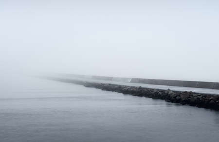 Panoramic view of Baltic sea from sandy shore. Promenade to the lighthouse, breakwaters. Thick white fog, mist. Waves, water splashes, long exposure. Seascape. Picturesque monochrome sceneryの写真素材