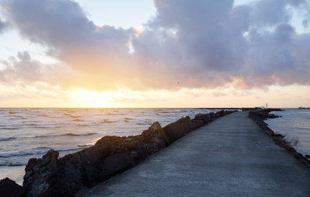 Old pier (walkway, promenade) to the lighthouse. Breakwaters close-up. Clear blue sky, pink sunset clouds, golden sunlight. Seascape, cloudscape. Baltic sea. Spring. Tourism, landmark, navigationの写真素材