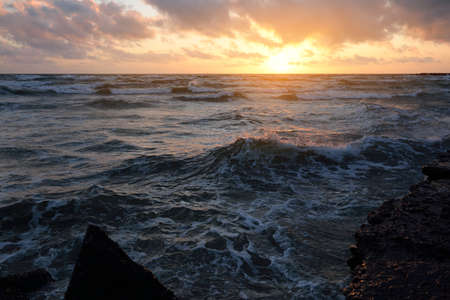 Baltic sea shore under a blue sky with glowing pink and golden sunset clouds. Rocky shore, breakwaters. Crashing waves. Latvia. Nature, environmental conservation, ecotourism. Picturesque sceneryの写真素材