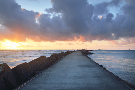Old pier (walkway, promenade) to the lighthouse. Breakwaters close-up. Clear blue sky, pink sunset...