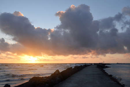 Old pier (walkway, promenade) to the lighthouse. Breakwaters close-up. Clear blue sky, pink sunset clouds, golden sunlight. Seascape, cloudscape. Baltic sea. Spring. Tourism, landmark, navigationの写真素材