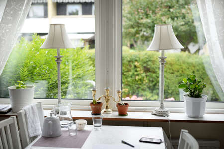 Interior of a classic swedish kitchen and dining room. White chairs, table, teapot, glass of water, teacup, candlestick, lamps. Green trees in the background. Stockholm, Sweden. House, home, decorの写真素材
