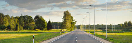 An empty highway (new asphalt road) through the green country fields and birch forest at sunset....