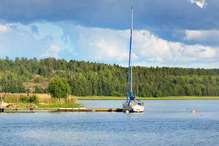 Sloop rigged yacht moored to a pier near the forest lakeshore. Balsta, MÃ¤laren lake, Sweden. Summer vacations, tourism, yachting, recreation, sport, leisure activity, lifestyleの写真素材