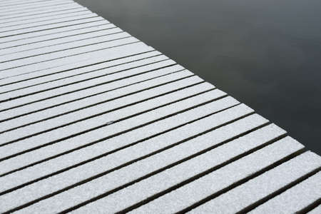 Snow-covered modern wooden pier (jetty, boardwalk) over the frozen river. Ice, frost, snow textures. Decoration, design, graphic resources, copy space, winter backgroundの写真素材
