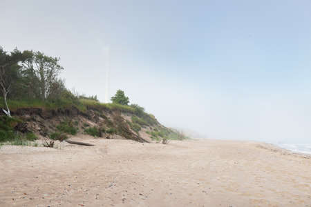 Baltic sea shore in a fog at sunrise. Panoramic view. Sand dunes, dune grass. Wind turbine generator in the background. Idyllic summer seascape. Power in nature, environment, ecology, renewable energyの写真素材