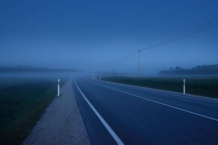 Country highway (asphalt road) in a fog at sunset. Moonrise, twilight, dramatic sky. Idyllic summer rural scene. Transportation, remote places, dangerous drivingの写真素材