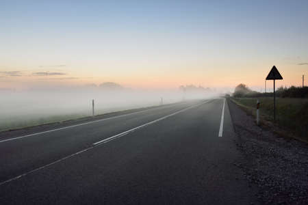 An empty highway (new asphalt road) through the fields and forest in a fog at sunset. Soft sunlight....