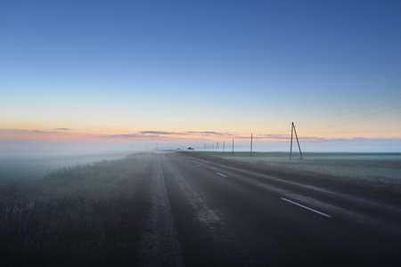 Country highway (asphalt road) in a fog at sunset. Electricity power line. Moonrise, twilight, dramatic sky, glowing golden clouds. Idyllic rural scene. Transportation, remote places, infrastructureの写真素材