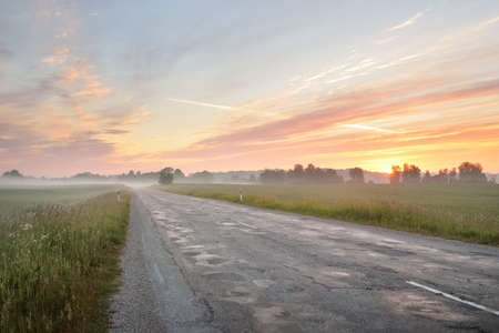 Country highway (old asphalt road) through the field and forest at sunrise. Soft sunlight, glowing pink and golden clouds. Vacations, logistics, remote placesの写真素材