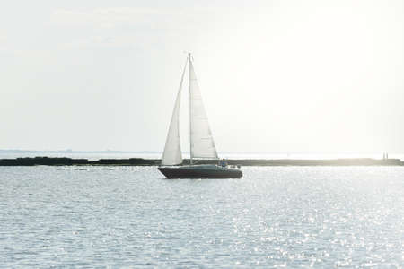 White sloop rigged yacht sailing in the Baltic sea at sunset. Clear sky after the storm, soft sunlight. Lighthouse. Transportation, travel, cruise, sport, recreation, leisure activity, racing, regattaの写真素材