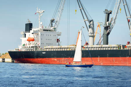 Sloop rigged yacht sailing in the Baltic sea. Cargo ship loading in port terminal in the background. Transportation, travel, cruise, sport, recreation, leisure activity, business, economy, industryの写真素材